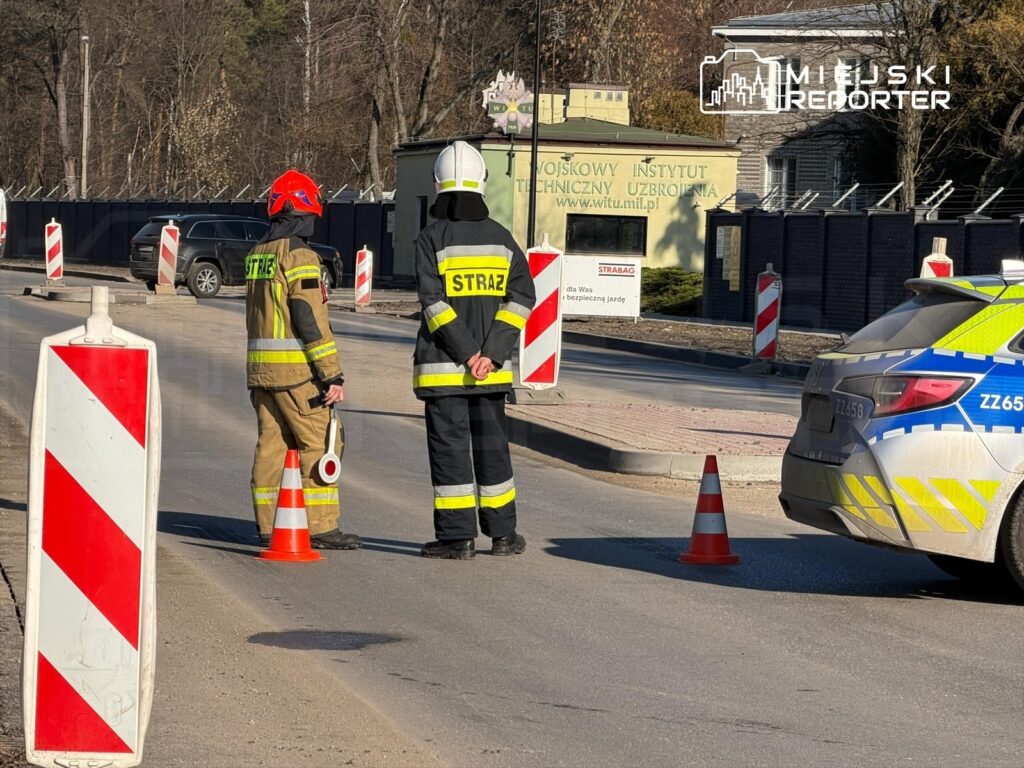 Strażacy i policjant stoją przy drodze, kontrolując ruch w rejonie budowy, otoczeni znakami drogowymi.