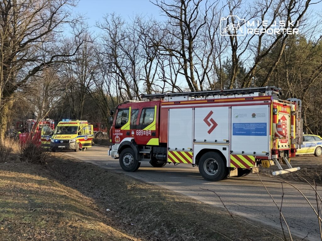 Wzdłuż drogi zatrzymane pojazdy służb ratunkowych, w tym wóz strażacki oraz ambulans, otoczone przez drzewa.
