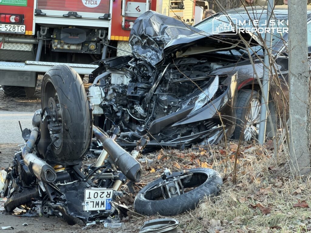 Zniszczony motocykl leży na poboczu drogi obok rozbitego samochodu, w tle widać woz strażacki.