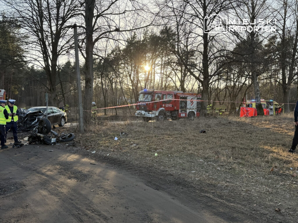 Na drodze leży wrak motocykla obok uszkodzonego samochodu; w tle strażacy oraz policjanci zabezpieczają miejsce zdarzenia.