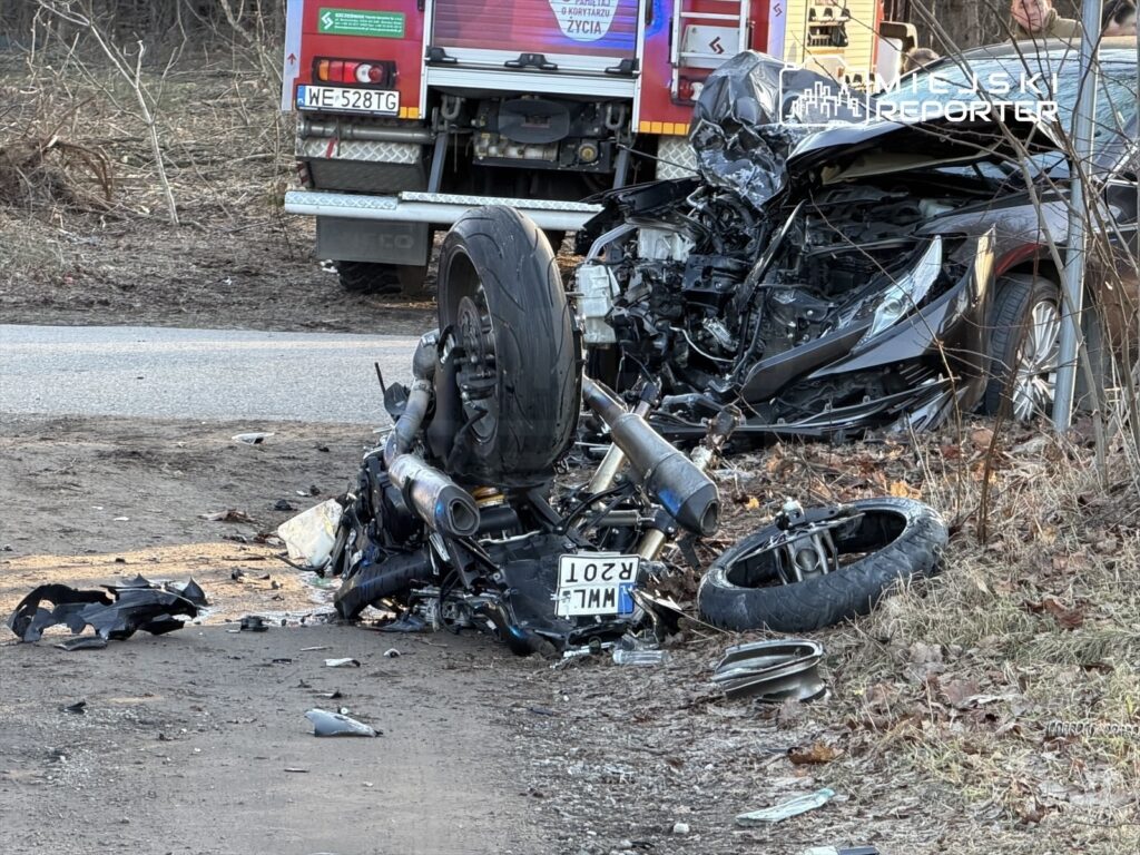 Zniszczony motocykl leży na boku obok uszkodzonego samochodu osobowego na poboczu drogi. W tle widoczny wóz strażacki.