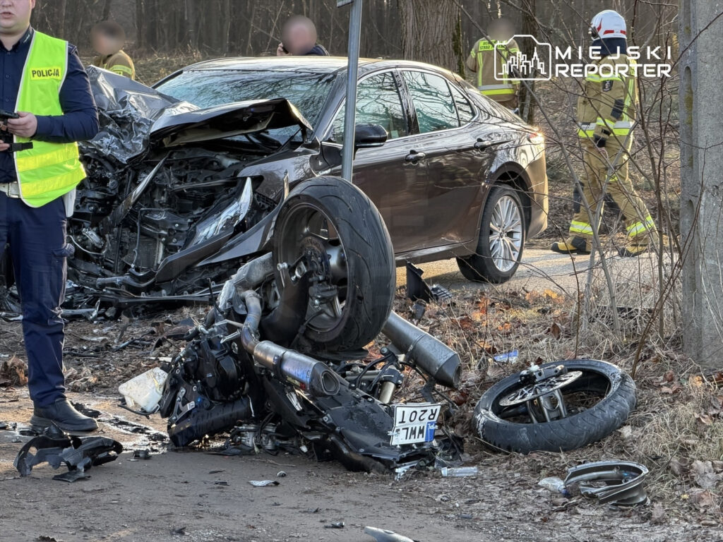 Mężczyzna w kamizelce odblaskowej rozmawia przez radio obok zniszczonego samochodu i motocykla na leśnej drodze.
