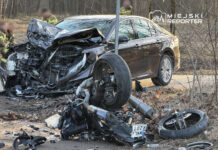 W wyniku wypadku drogowego zniszczony samochód osobowy stoi na poboczu, obok leży przewrócony motocykl, a strażacy pracują na miejscu.