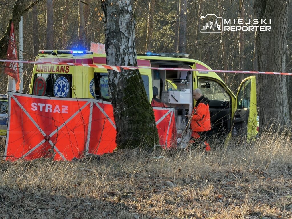 Ratownik w pomarańczowej kurtce przygotowuje sprzęt w otwartej karetce, otoczonej taśmą zabezpieczającą w lesie.