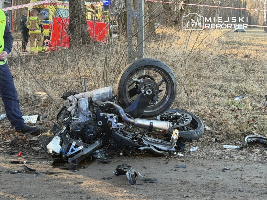 Motocykl leżący na boku z uszkodzoną konstrukcją na poboczu drogi, obok policjant i zespół ratunkowy.