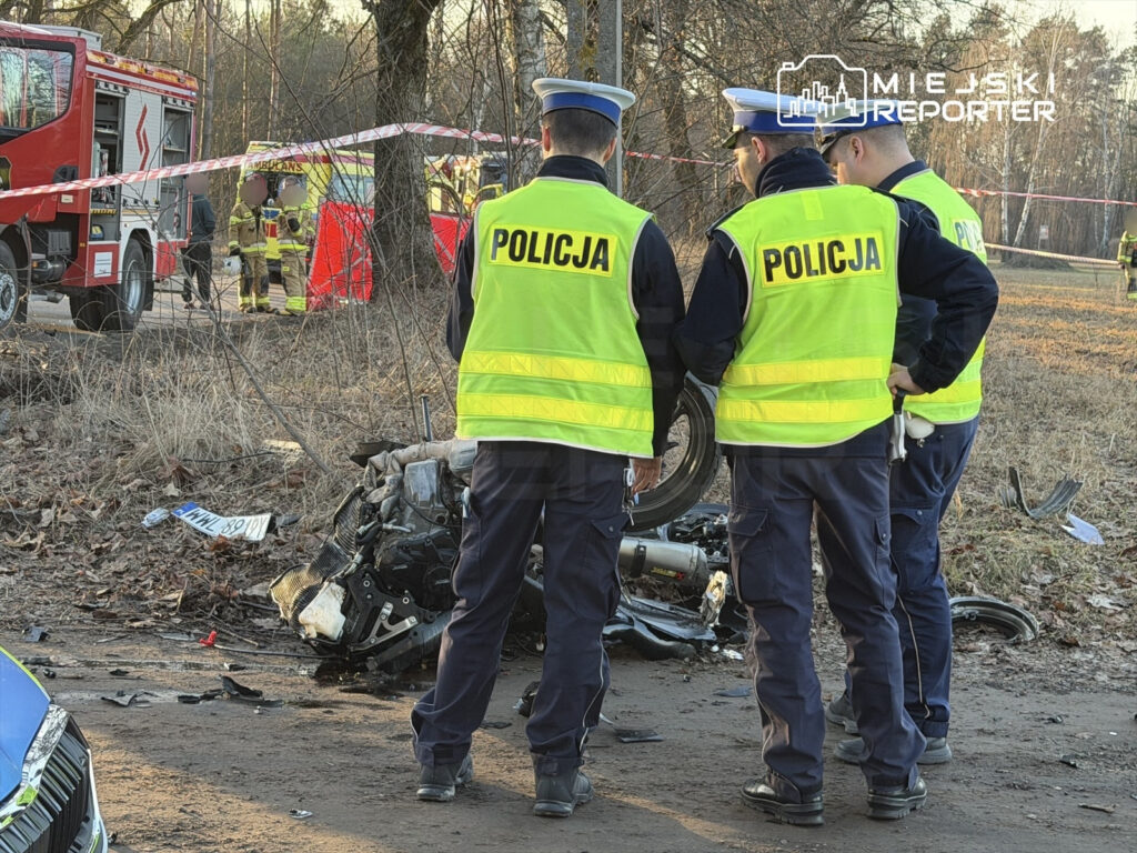 Grupa policjantów w kamizelkach odblaskowych obserwuje wrak motocykla na poboczu drogi, obok działa ekipa strażaków.