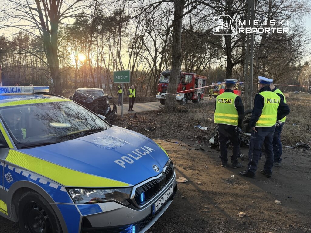 Policyjny radiowóz z włączonymi sygnałami na drodze, obok grupy policjantów badających miejsce wypadku. W tle widać uszkodzony samochód oraz woz strażacki.