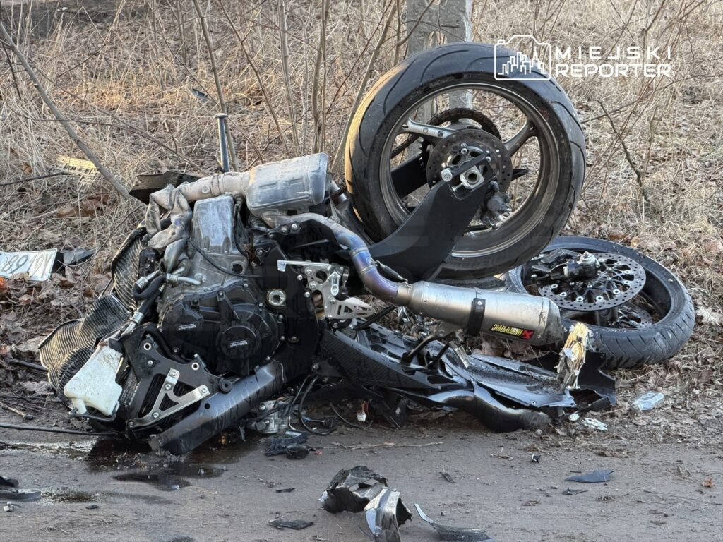 Zniszczony motocykl leży na ziemi w zaroślach, obok widoczne są odłamki części oraz koło.