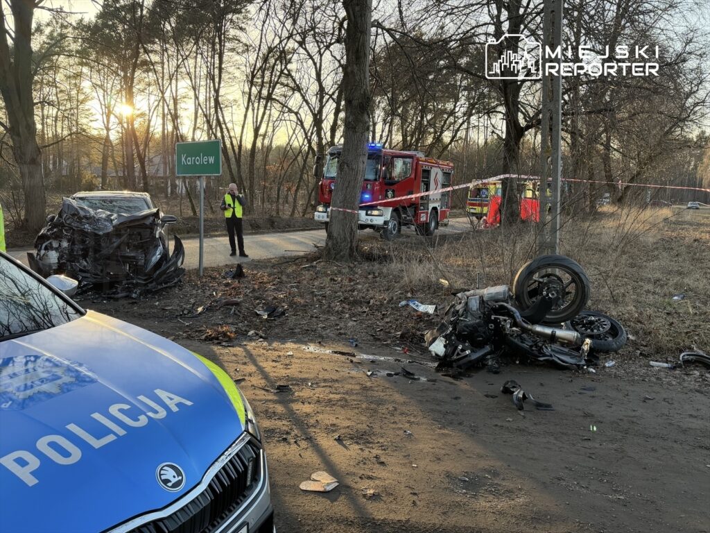 W miejscu wypadku drogowego widać uszkodzony samochód oraz rozbite moto, a w tle pracują strażacy i policjant telefonujący.