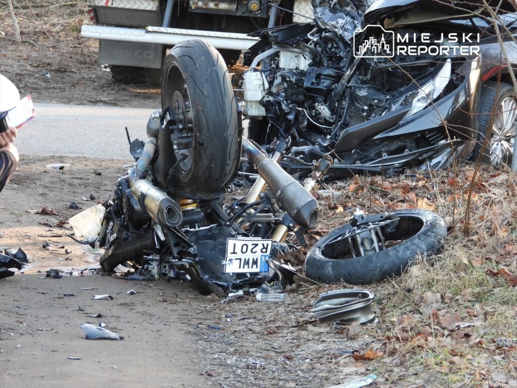 Motocykl leżący na boku obok zniszczonego samochodu na poboczu drogi, otoczony fragmentami wraku.