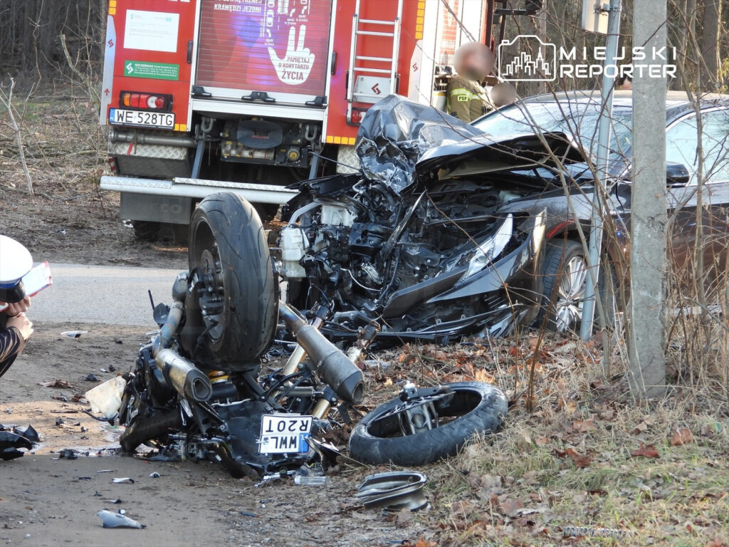 W wyniku wypadku zniszczony motocykl leży na poboczu drogi, obok stoi straż pożarna, a funkcjonariusz dokumentuje miejsce zdarzenia.