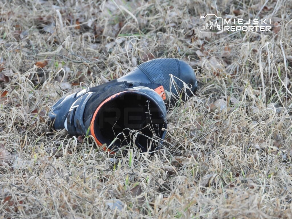 Czarny but sportowy leży na wyschniętej trawie, z odkrytą podeszwą i pomarańczowym detalem.