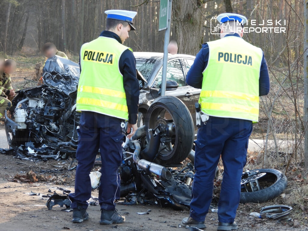 Policjanci w odblaskowych kamizelkach badają miejsce wypadku drogowego, obok zniszczonego motocykla i samochodu.