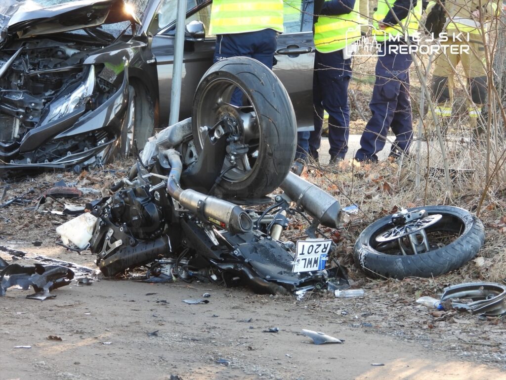 Zniszczony motocykl leży na ziemi obok uszkodzonego samochodu, a w tle interweniują ratownicy.