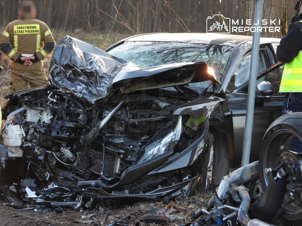 Zniszczony samochód osobowy z uszkodzoną przednią częścią stoi przy drodze, obok pracują strażacy i policjanci.