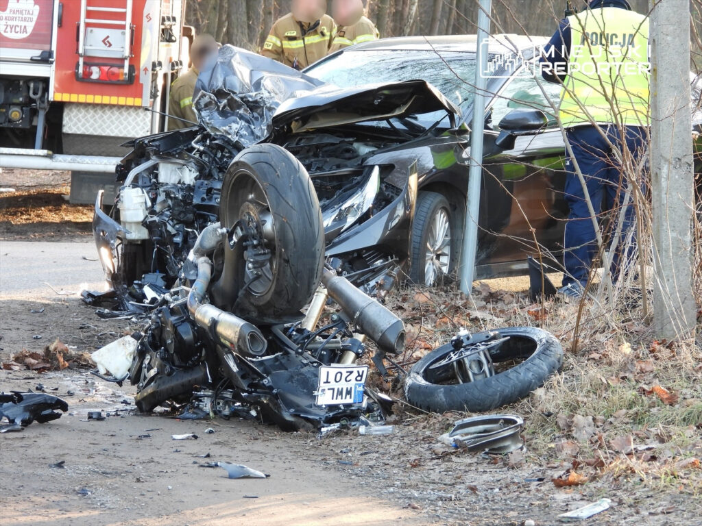 Zniszczony motocykl leży obok rozbitego samochodu na poboczu drogi, w tle strażacy i policjanci interweniują.