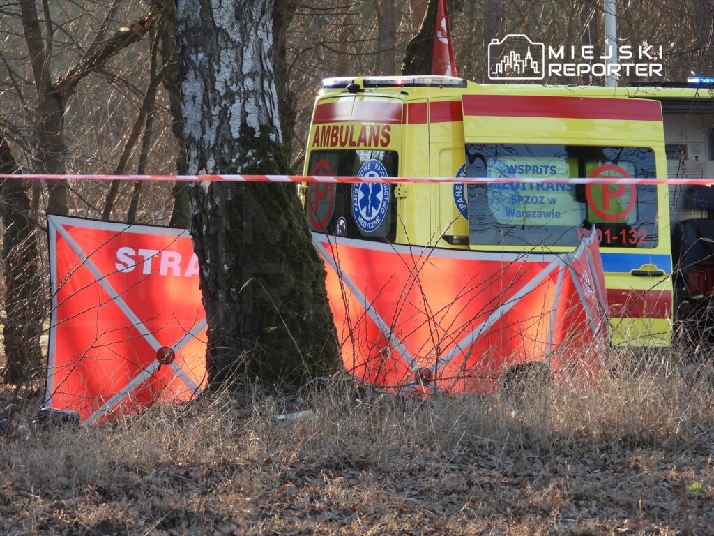 Zespół ratownictwa medycznego zaparkowany obok drzewa, otoczony czerwonymi oznaczeniami i taśmą zabezpieczającą.