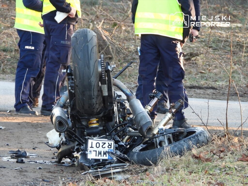 Motocykl leżący na boku z uszkodzeniami, obok policjanci w kamizelkach odblaskowych prowadzący dochodzenie.