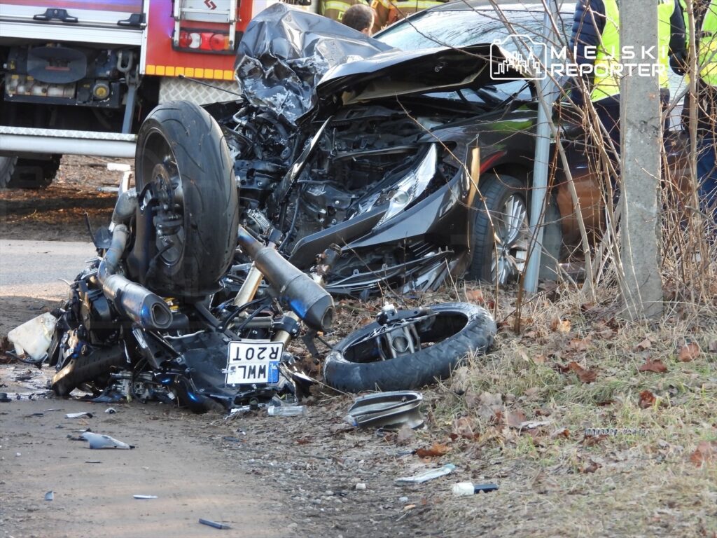 Zniszczony motocykl leży na poboczu drogi obok uszkodzonego samochodu, w tle widać strażaków oraz pojazdy ratunkowe.