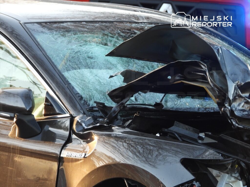 Uszkodzony samochód osobowy z rozbitą szybą przednią, stojący na drodze, obok widać pojazdy policyjne.