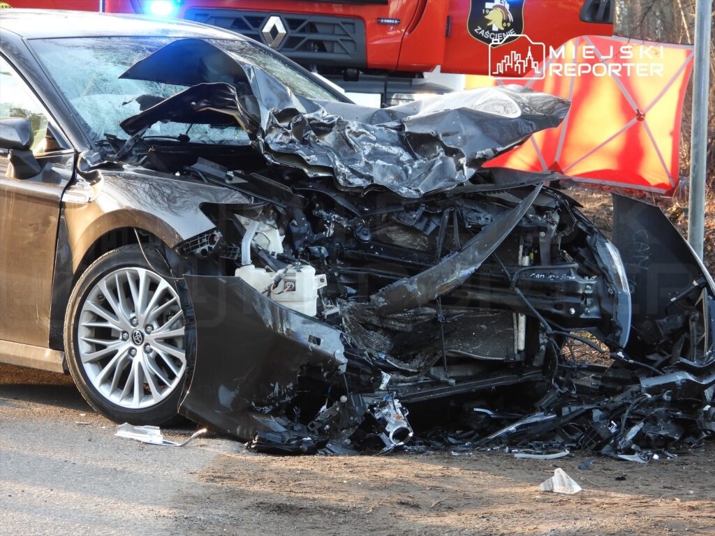 Zniszczony samochód osobowy leży na poboczu drogi, obok widać wóz strażacki oraz elementy akcji ratunkowej.