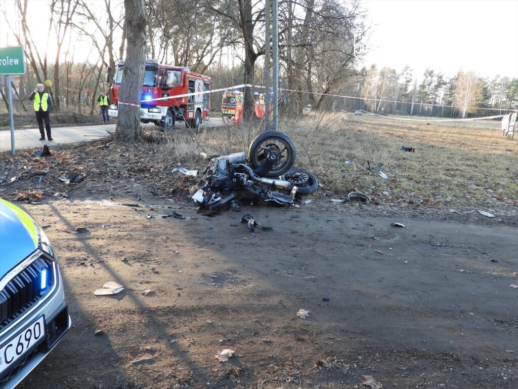 Motocykl leżący na boku na poboczu drogi, obok widoczne resztki wypadku oraz pojazdy służb ratunkowych.