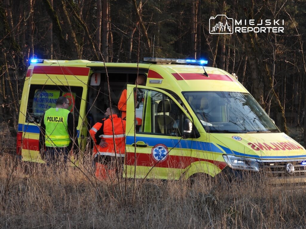 Ratownicy medyczni w pomarańczowych strojach rozmawiają z policjantem w pobliżu karetki w lesie.