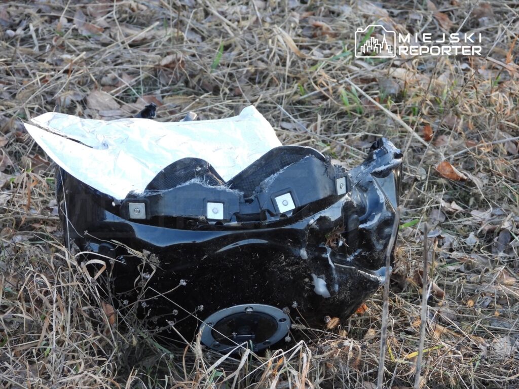Zniszczony element pojazdu leży na trawie, otoczony suchymi liśćmi i gałązkami.