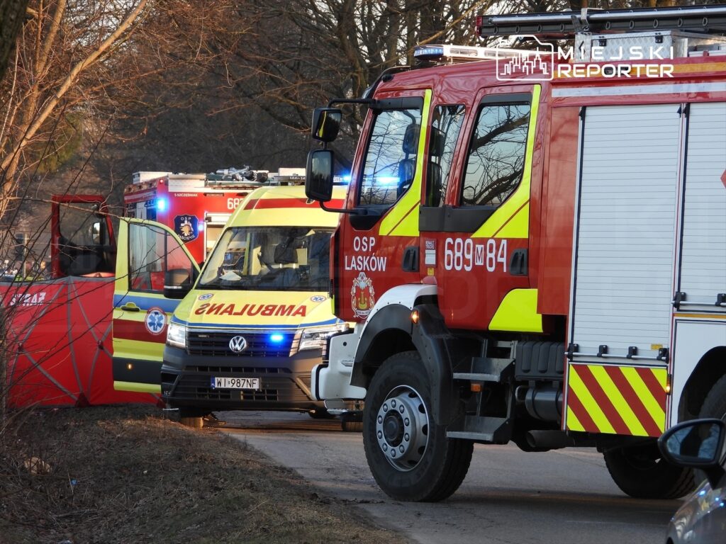 Zespół ratunkowy z ambulansu i wozu strażackiego na miejscu zdarzenia w otoczeniu drzew.