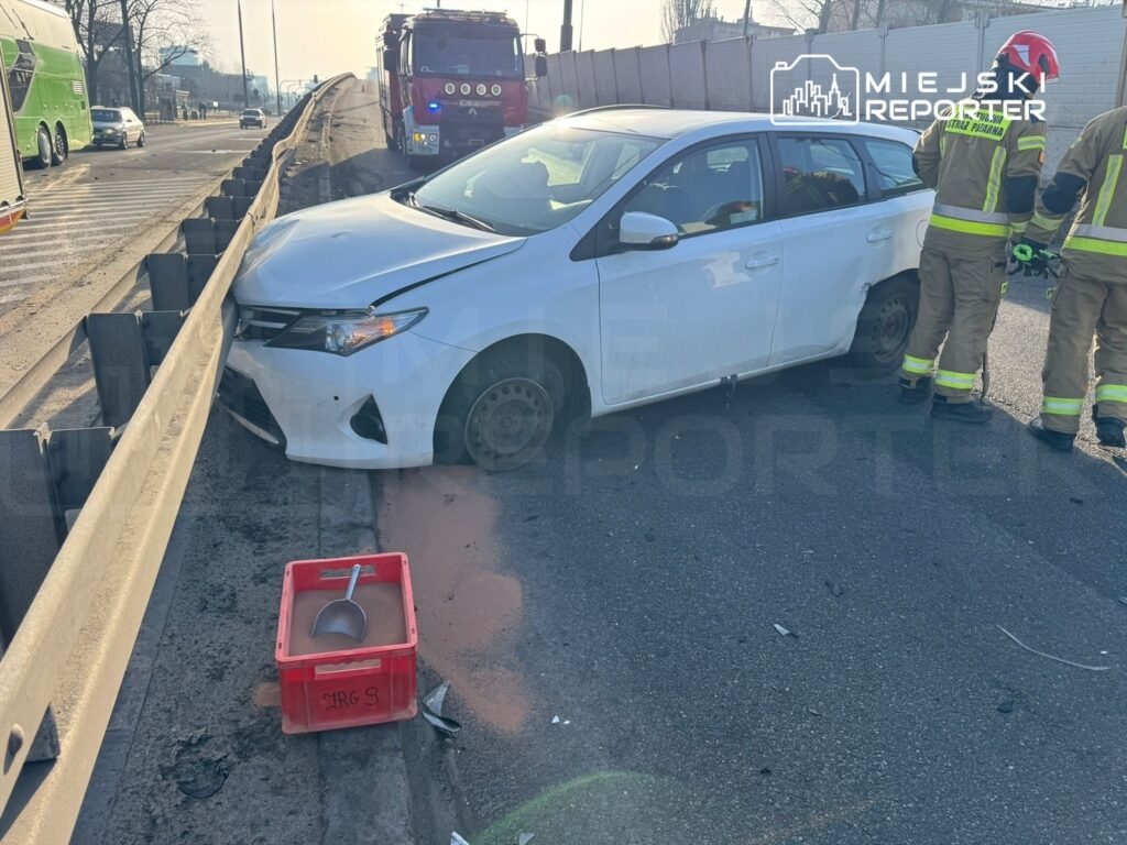 Biały samochód osobowy uderzony w barierkę, obok strażacy pracujący przy zdarzeniu drogowym na ruchliwej ulicy.