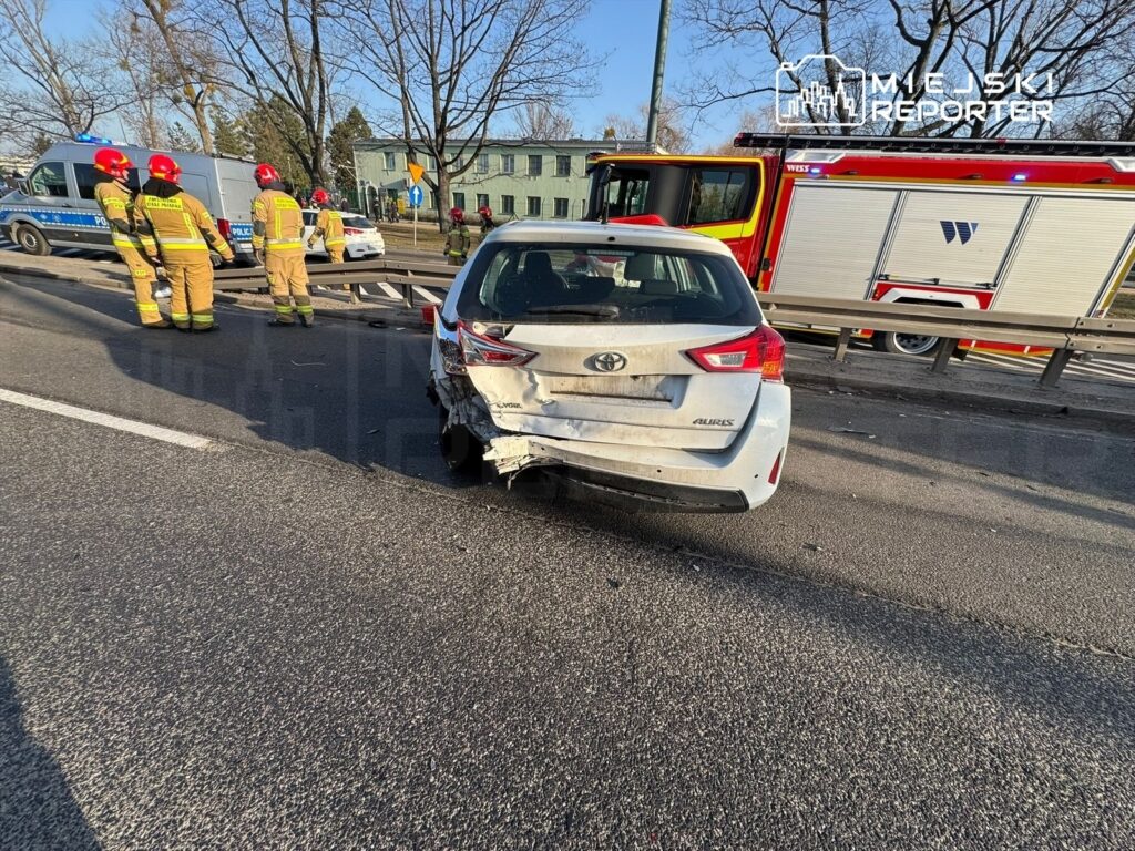 Wielu strażaków w mundurach pracuje przy uszkodzonym samochodzie osobowym na drodze, w tle widać woz strażacki.
