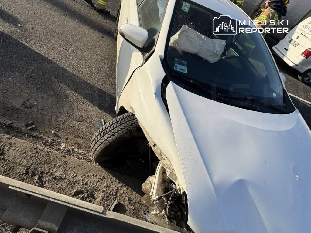 Wybite koło białego samochodu osobowego, uszkodzone nadwozie przy krawężniku, w tle interwencja służb ratunkowych.