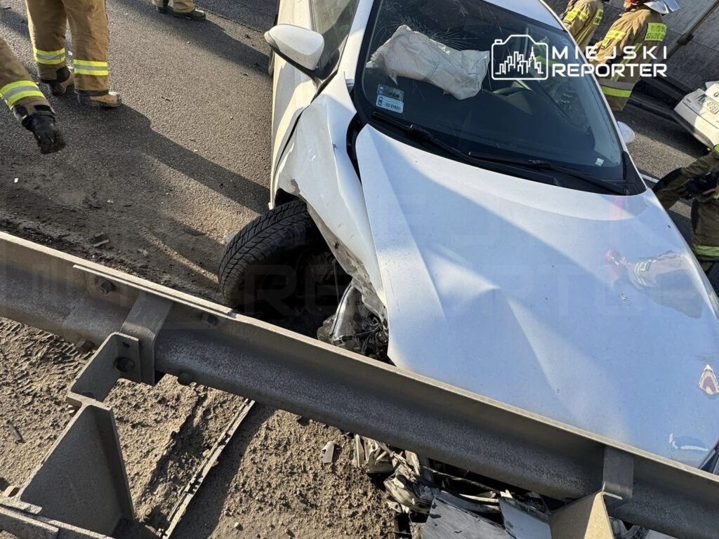 Biały samochód osobowy uderzył w barierkę drogową, obok pracują strażacy w mundurach.