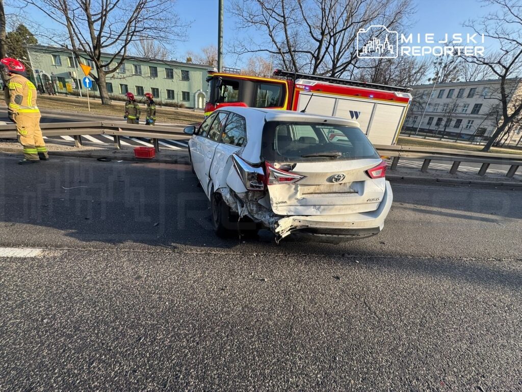 Uszkodzony samochód osobowy stoi obok bariery drogowej, obok niego pracują strażacy w mundurach.