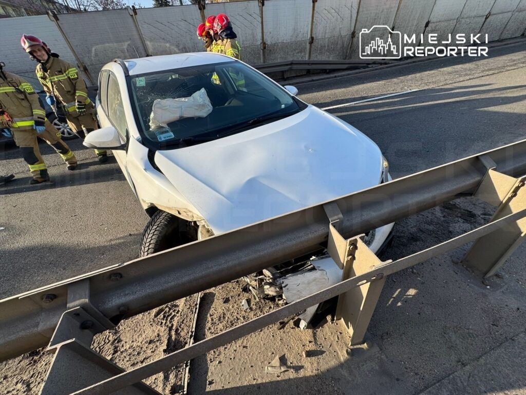 Biały samochód osobowy uderzył w barierkę, obok stoją strażacy w mundurach, oceniając sytuację na drodze.