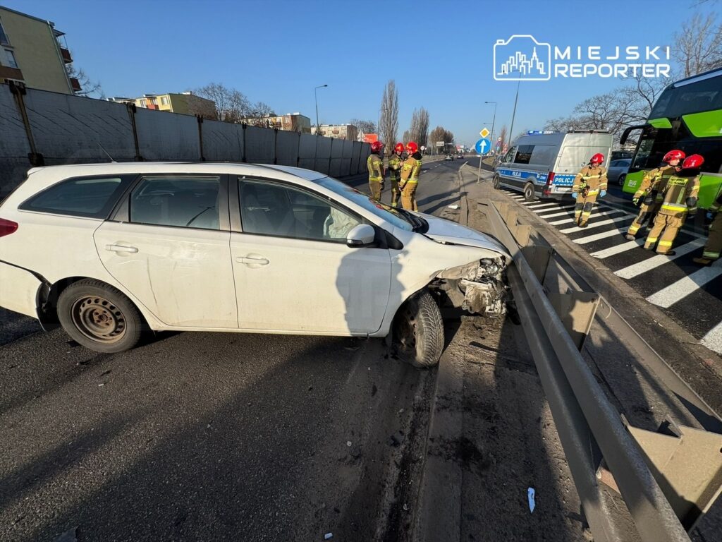 Biały samochód osobowy uderzony w barierkę drogową, obok grupka strażaków i radiowóz policyjny na skrzyżowaniu.