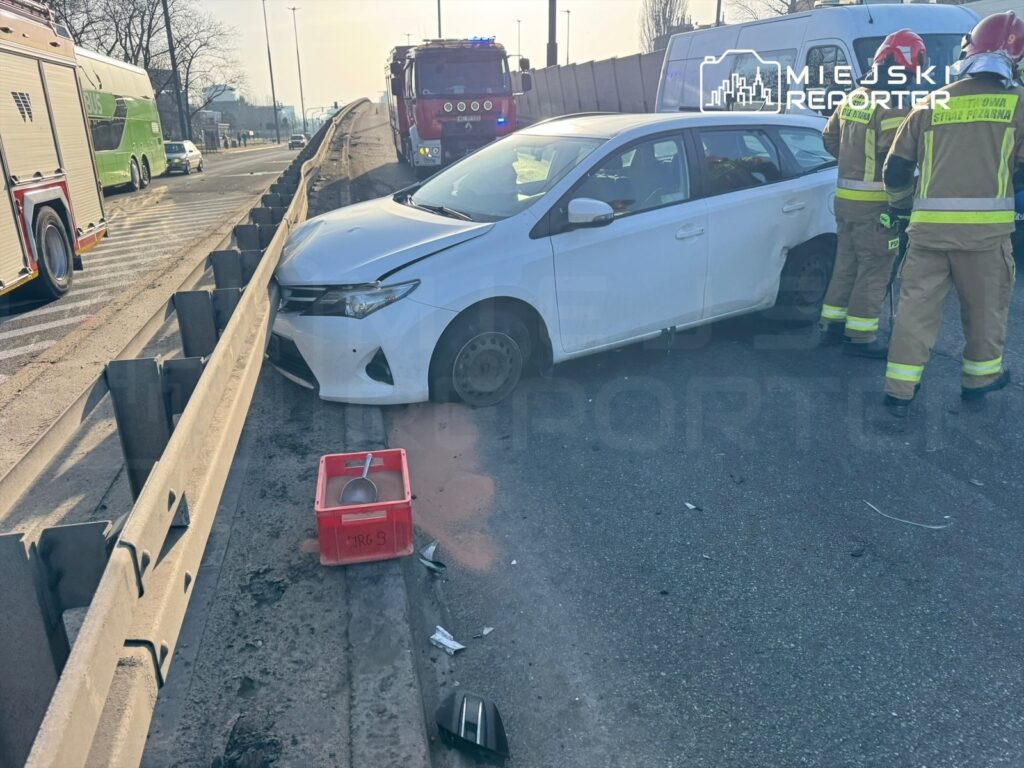 Biały samochód osobowy uderzony w barierę drogową, obok strażacy w uniformach interweniujący na miejscu zdarzenia.