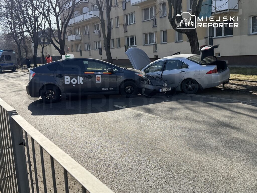 Czarny samochód osobowy z logo Bolt zderzył się z szarym autem na ulicy, obok stoją radiowóz i świadkowie.