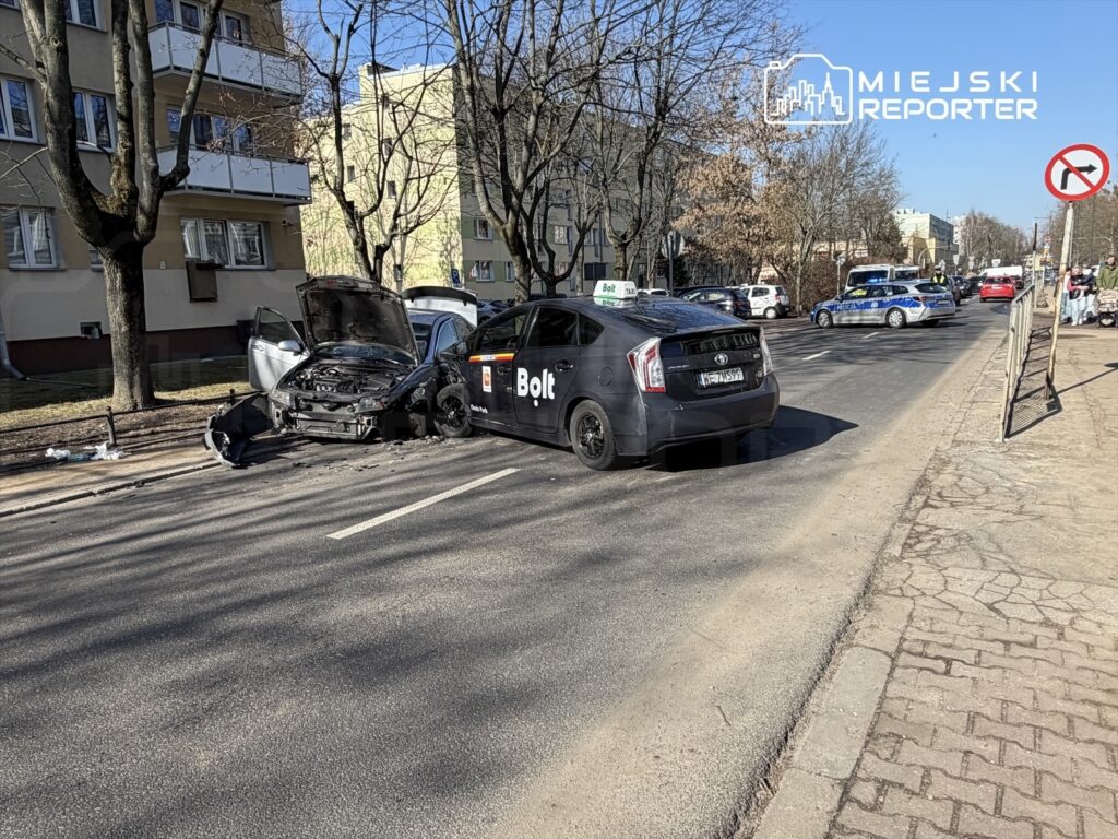 Zderzenie dwóch samochodów na ulicy; jeden z pojazdów ma otwartą maskę, a w tle widoczne są inne samochody oraz budynki mieszkalne.