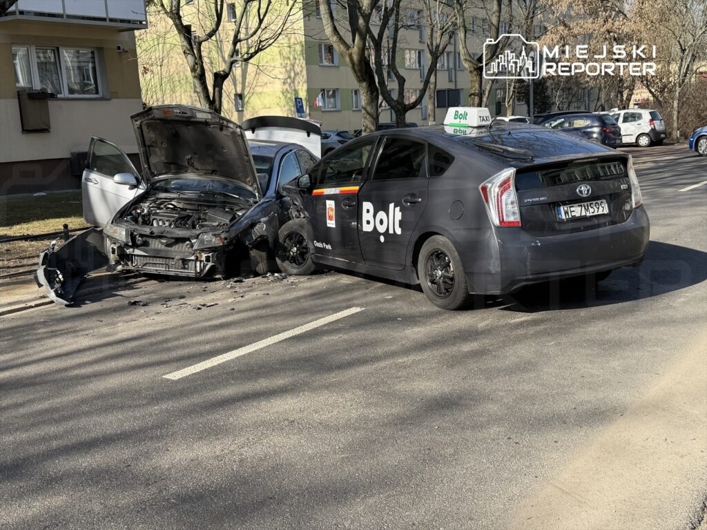 Zniszczony samochód osobowy z otwartą maską stoi na poboczu obok taksówki Bolt w miejskim otoczeniu.
