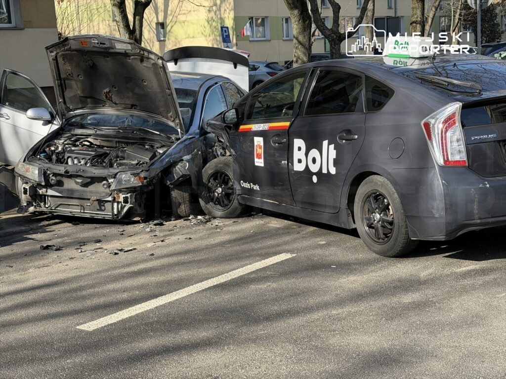 Uszkodzony samochód osobowy z otwartą maską stoi obok taksówki Bolt, obie pojazdy zaparkowane na ulicy.