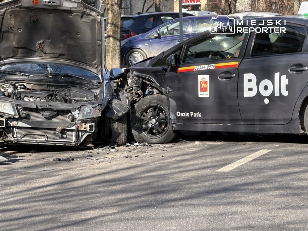 Dwa uszkodzone samochody po zderzeniu na parkingu, w tym czarny samochód z logo Bolt i pojazd z uniesioną maską.