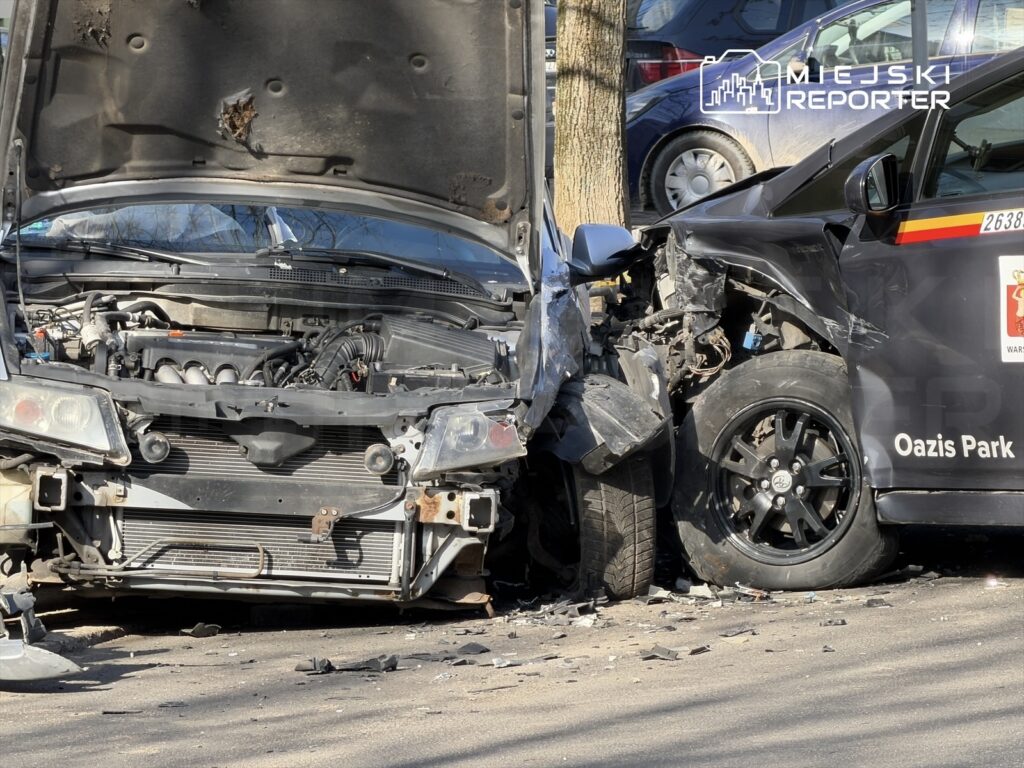 Zderzenie dwóch samochodów na ulicy, jeden z pojazdów ma otwartą maskę, widoczne uszkodzenia.