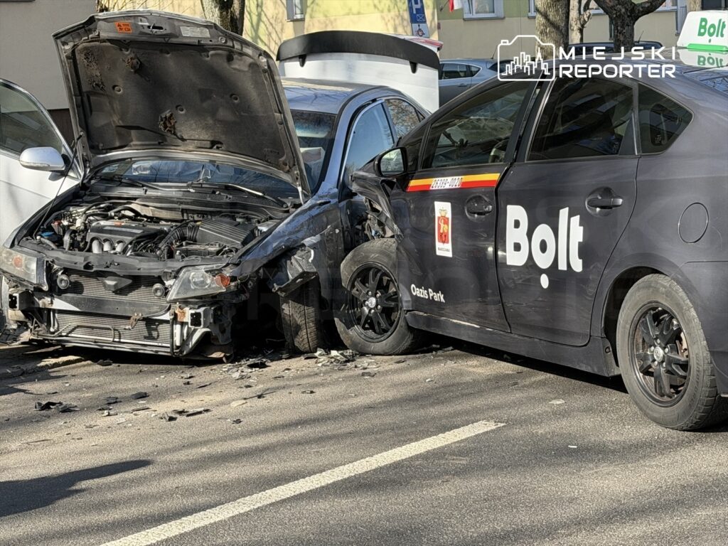 Dwa uszkodzone samochody, w tym czarny pojazd z logo Bolt, stoją na ulicy po kolizji. Otwarta maska jednego z aut.