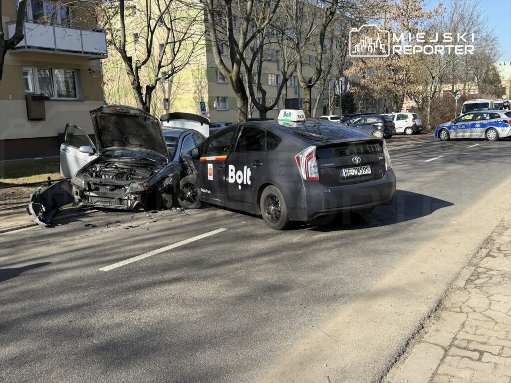 Zderzenie dwóch samochodów na ulicy, jeden z pojazdów ma uszkodzoną przednią część, a drugi stoi obok.