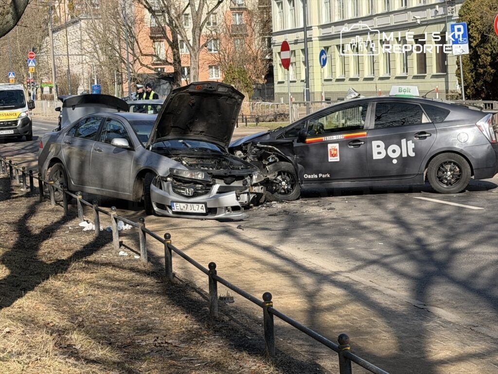 Dwa uszkodzone samochody po zderzeniu na ulicy, jeden z otwartą maską, w tle radiowóz i przechodnie.