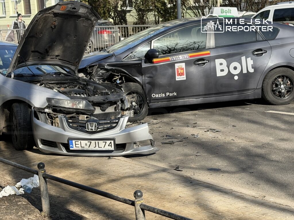 Uszkodzony srebrny samochód osobowy z otwartą maską stoi obok czarnego auta z logo Bolt, w tle widać chodnik i drzewa.
