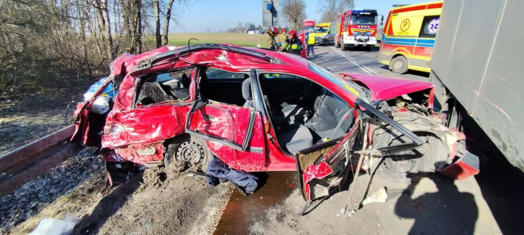 Rozbity czerwony samochód osobowy z wygiętą karoserią stoi obok drogi, w tle widoczni ratownicy i wozy strażackie.