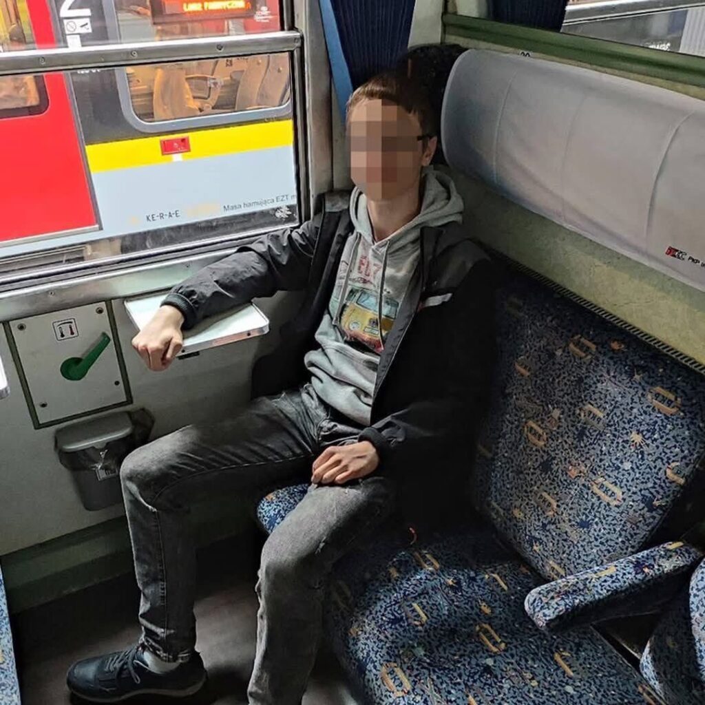 Młody mężczyzna w szarej bluzie siedzi na niebieskiej kanapie w pociągu, obok okna z widokiem na inny pociąg.