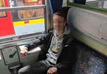Młody mężczyzna w szarej bluzie siedzi na niebieskiej kanapie w pociągu, obok okna z widokiem na inny pociąg.